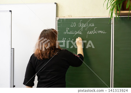 A dedicated teacher writes complex math exercises on a green chalkboard in a primary school classroom to foster academic focus and engagement for young elementary level students A dedicated teacher writes complex math exercises on a green chalkboard in a primary school classroom to foster academic focus and engagement for young elementary level students 141313924