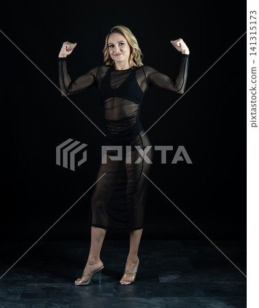 Young blonde dancer wearing transparent black dress and high heels posing in studio with black background Young blonde dancer wearing transparent black dress and high heels posing in studio with black background 141315173
