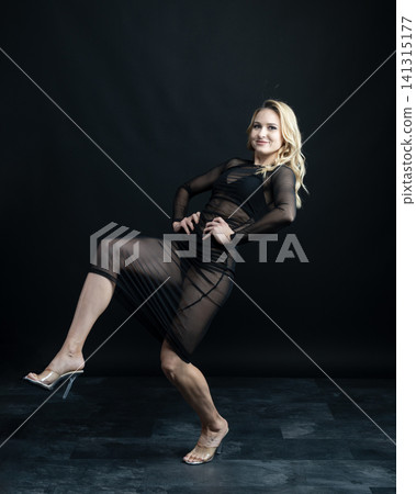 Young blonde dancer wearing transparent black dress and high heels posing in studio with black background Young blonde dancer wearing transparent black dress and high heels posing in studio with black background 141315177