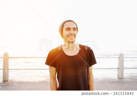 Man, smile and promenade at beach in sunset with streetwear, beanie and vacation by ocean. Person, outdoor and happy by sea, waves and boardwalk on holiday with nature, sunshine and thinking in Italy Man, smile and promenade at beach in sunset with streetwear, beanie and vacation by ocean. Person, outdoor and happy by sea, waves and boardwalk on holiday with nature, sunshine and thinking in Italy 141315842