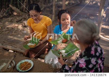 Two women and young girl sharing homemade meal on bamboo platform outdoors with banana leaf plates during relaxed community gathering in rustic rural setting with warm natural sunlight Two women and young girl sharing homemade meal on bamboo platform outdoors with banana leaf plates during relaxed community gathering in rustic rural setting with warm natural sunlight 141316674