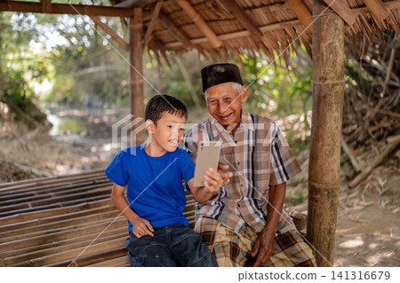 Smiling boy and elderly man sharing smartphone selfie on bamboo bench beneath rustic shelter, warm daylight in countryside scene showing connection across generations and relaxed outdoor togetherness Smiling boy and elderly man sharing smartphone selfie on bamboo bench beneath rustic shelter, warm daylight in countryside scene showing connection across generations and relaxed outdoor togetherness 141316679
