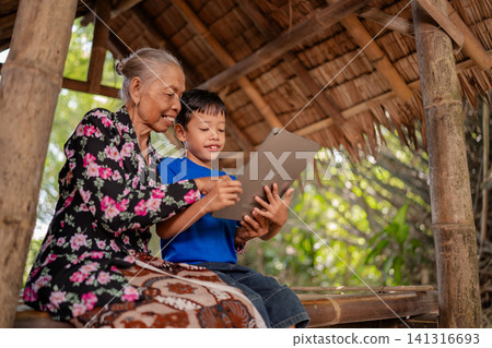 Elderly woman and young boy sharing tablet screen inside rustic bamboo shelter, smiling together in warm daylight with family learning, intergenerational bonding, and rural lifestyle atmosphere 141316693