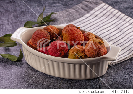Fresh lychee fruits in white ceramic dish with green leaves on textured gray surface 141317312