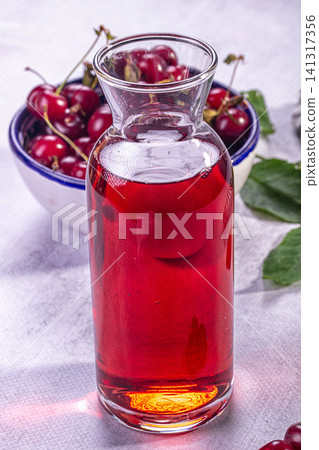 Glass carafe filled with red cherry juice beside bowl of fresh cherries on light surface 141317356