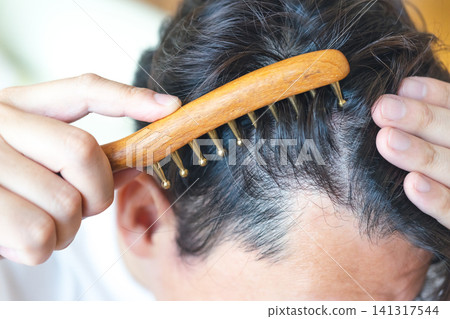 A man in his 40s massaging his scalp with a comb-shaped gua sha tool. A man in his 40s massaging his scalp with a comb-shaped gua sha tool. 141317544