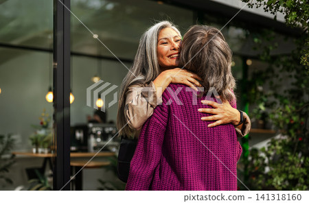 Genuine Connection Captured as a Smiling Silver Haired Woman Hugs a Friend Closely Outside a Modern Glass Cafe Framed by Green Foliage 141318160