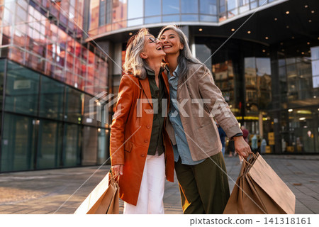 Joyful Mature Women Friends Sharing a Playful Cheek Kiss Outside a Modern Glass Shopping Center Holding Kraft Bags in Earth Tone Outfits 141318161