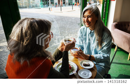 Joyful Silver Haired Woman Toasting Coffee Cups with a Friend Inside a Bright Urban Cafe by a Street Window Joyful Silver Haired Woman Toasting Coffee Cups with a Friend Inside a Bright Urban Cafe by a Street Window 141318162