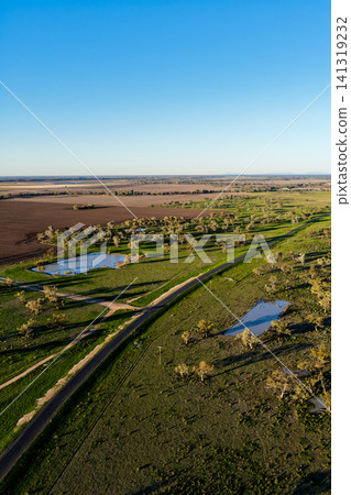 Aerial vertical scene showcasing cultivated farmland with a rural dirt road connecting to a main road, scattered farm dams and a lush green landscape near Pilliga, highlighting the striking patterns Aerial vertical scene showcasing cultivated farmland with a rural dirt road connecting to a main road, scattered farm dams and a lush green landscape near Pilliga, highlighting the striking patterns 141319232