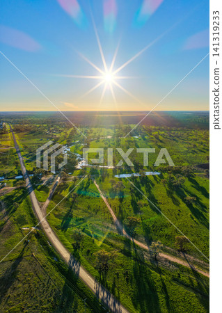 Aerial vertical scene showcasing a lush green rural landscape near Pilliga, featuring a dirt road connecting to a main road, scattered farm dams and the sun creating a striking sunstar Aerial vertical scene showcasing a lush green rural landscape near Pilliga, featuring a dirt road connecting to a main road, scattered farm dams and the sun creating a striking sunstar 141319233