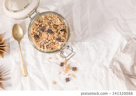 Crunchy granola with dark chocolate chunks in a glass jar on a white cloth for a healthy and nutritious morning breakfast meal Crunchy granola with dark chocolate chunks in a glass jar on a white cloth for a healthy and nutritious morning breakfast meal 141320088