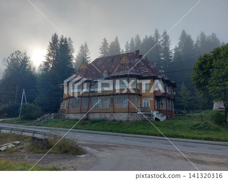 Atmospheric old wooden mountain house surrounded by dense pine forest on a misty morning. Historic rural architecture, soft sunlight through fog, peaceful countryside landscape with copy space for Atmospheric old wooden mountain house surrounded by dense pine forest on a misty morning. Historic rural architecture, soft sunlight through fog, peaceful countryside landscape with copy space for 141320316
