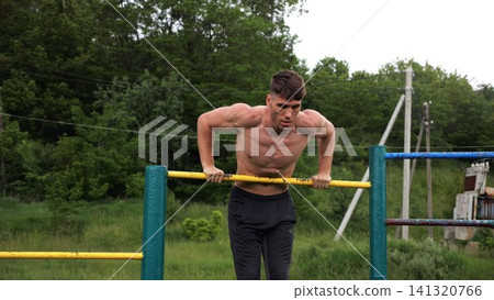 Hardy shirtless sportsman doing push ups on horizontal bar at nature. Strong and muscular guy training at sports ground. Young athletic guy exercising outdoor. Concept of active and healthy lifestyle Hardy shirtless sportsman doing push ups on horizontal bar at nature. Strong and muscular guy training at sports ground. Young athletic guy exercising outdoor. Concept of active and healthy lifestyle 141320766