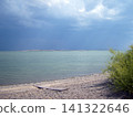 Dark, ominous storm clouds gather over a vast, calm lake. The pebbly shoreline features a single wooden plank and a vibrant green bush, contrasting with the moody, dramatic atmosphere of the landscape 141322646