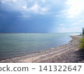 A dramatic landscape featuring a calm lake alongside a curving rocky shoreline. A dark, ominous storm cloud approaches from the left, contrasting sharply with the bright, sunlit sky on the right 141322647
