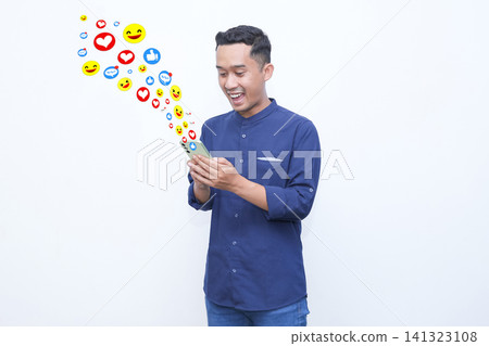 Young man smiles while holding smartphone. Colorful emojis float from his device. Joyful expressions radiate from his face. Ideal for social media or digital communication themes Young man smiles while holding smartphone. Colorful emojis float from his device. Joyful expressions radiate from his face. Ideal for social media or digital communication themes 141323108