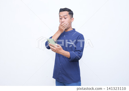 Man holds phone, hand covers mouth in shock. His eyes widen, eyebrows raised. White background isolates the emotion clearly. Ideal for stories of surprise or unexpected news. 141323110