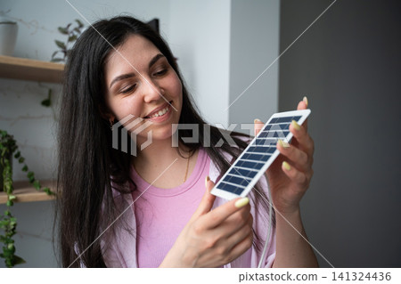 Young woman holding a portable solar panel indoors with a smile. Renewable energy education and sustainability technology concept Young woman holding a portable solar panel indoors with a smile. Renewable energy education and sustainability technology concept 141324436