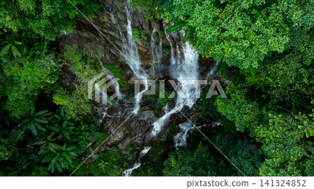 Aerial view of deep tropical rainforest with waterfall surrounded by lush tropical forest and towering cliffs streams of water into deep canyon, Lush green trees in forest with natural stream. 141324852