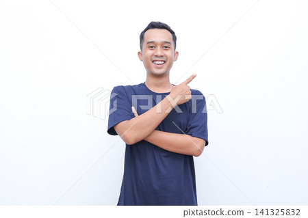 Young man smiles, pointing with confident gesture. Crossed arms show relaxed, friendly attitude. Bright background emphasizes his cheerful expression. Ideal for business, promotional 141325832