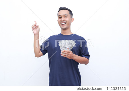 Young man smiles broadly, holding cash. He points upward with one hand. White background keeps focus on him. His expression shows joy and excitement. Ideal for financial success or achievement themes Young man smiles broadly, holding cash. He points upward with one hand. White background keeps focus on him. His expression shows joy and excitement. Ideal for financial success or achievement themes 141325833