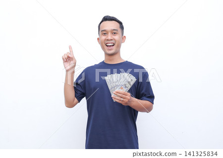 Young man smiles broadly, holding cash. He points upward with one hand. White background keeps focus on him. His expression shows joy and excitement. Ideal for financial success or achievement themes Young man smiles broadly, holding cash. He points upward with one hand. White background keeps focus on him. His expression shows joy and excitement. Ideal for financial success or achievement themes 141325834