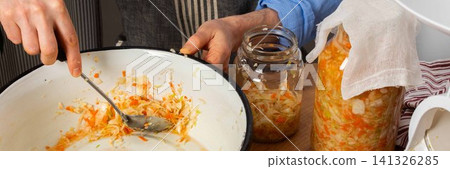 In a kitchen, a 53-year-old man works on fermenting cabbage and carrots. He carefully transfers the mixture into a glass jar for storage and fermentation, banner In a kitchen, a 53-year-old man works on fermenting cabbage and carrots. He carefully transfers the mixture into a glass jar for storage and fermentation, banner 141326285