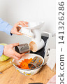 A 53-year-old man is in his kitchen preparing cabbage and carrots for fermentation. He uses a machine to shred the vegetables before placing them in a bowl 141326286