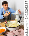 A 53-year-old man prepares cabbage and carrots for fermentation in his kitchen. He uses a machine to shred the vegetables while surrounded by kitchen tools and ingredients 141326287