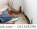 Person wearing blue head covering, safety goggles, and orange gloves applies cement to an apartment balcony wall. Renovation work improves the open outdoor space, focusing on corner repairs 141326290
