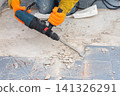 Person in orange gloves and work clothes uses a hammer drill to chip old grey tiles from an open apartment balcony floor. The tool breaks the surface, creating dust and tile debris 141326291