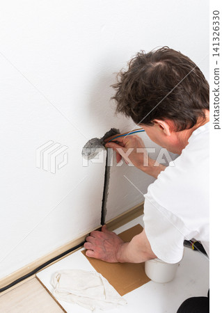 Worker runs new electrical wires into a cutout in a white apartment wall. This task prepares for the installation of an electrical outlet near the baseboard Worker runs new electrical wires into a cutout in a white apartment wall. This task prepares for the installation of an electrical outlet near the baseboard 141326330