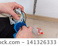 Bare hands connect blue, brown, and green electrical wires to a new wall socket mechanism. This installation occurs inside an apartment, preparing the wall for a functional power outlet 141326333