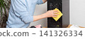 A 53-year-old man uses a cloth to clean dust from a computer system unit. He works at a wooden table in a room with plants nearby. Tools are set out for the task, banner 141326334