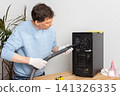 A 53-year-old man cleans dust from a computer system unit with a vacuum cleaner in a tidy room. He carefully removes dirt to keep the device running well 141326335