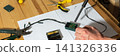 A man works on fixing a broken Christmas tree garland by soldering wires together. He uses tools and parts on a wooden table, focusing on the repair process during the holiday season 141326336