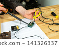 A man works on fixing a broken Christmas tree garland by soldering wires together. He uses tools and parts on a wooden table, focusing on the repair process during the holiday season 141326337