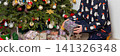 A 53-year-old man kneels by a Christmas tree in his home on Christmas Day. He examines a gift carefully as holiday decorations surround him. The moment is filled with family joy and cheer 141326348