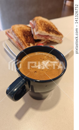 Black coffee served in a ceramic mug on a table, with toasted sandwiches arranged on a plate in the background, creating a casual dining atmosphere Black coffee served in a ceramic mug on a table, with toasted sandwiches arranged on a plate in the background, creating a casual dining atmosphere 141326752