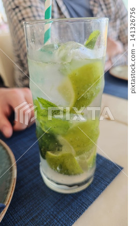 Mojito cocktail featuring fresh mint and lime slices in a tall glass, placed on a blue tablecloth with a hand visible in the background Mojito cocktail featuring fresh mint and lime slices in a tall glass, placed on a blue tablecloth with a hand visible in the background 141326756