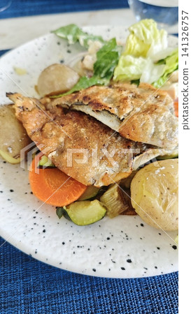 Grilled fish fillet served on a white plate with assorted vegetables including carrots, zucchini, and potatoes alongside a fresh green salad 141326757