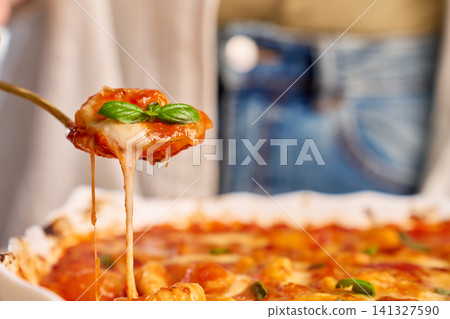 Gnocchi in tomato sauce served on spoon with cheese stretching from baking dish and basil on top 141327590