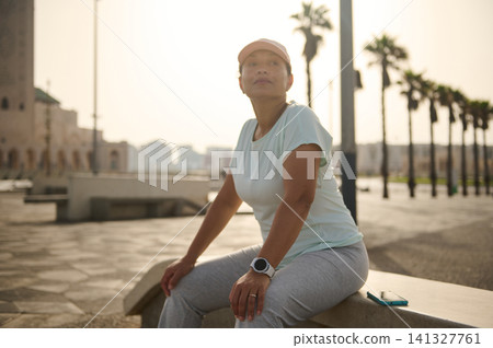 Middle-aged woman resting after morning run on urban promenade with palm trees and smartwatch Middle-aged woman resting after morning run on urban promenade with palm trees and smartwatch 141327761