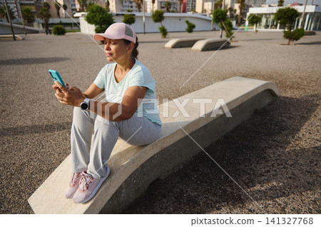 Woman resting on concrete bench after outdoor workout checking smartphone and smartwatch Woman resting on concrete bench after outdoor workout checking smartphone and smartwatch 141327768