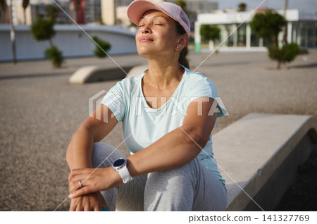 Woman relaxing after outdoor workout, resting in the sun with cap and smartwatch 141327769