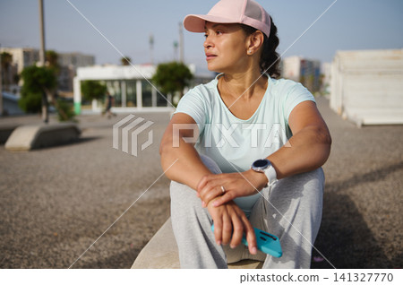 Woman resting after outdoor workout wearing cap and smart-watch in urban park Woman resting after outdoor workout wearing cap and smart-watch in urban park 141327770