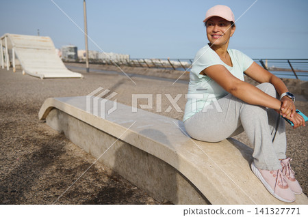 Smiling woman resting after seaside jog in activewear and cap on coastal promenade Smiling woman resting after seaside jog in activewear and cap on coastal promenade 141327771