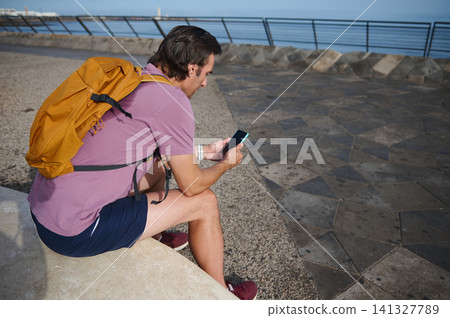 Man with yellow backpack sitting by the seaside checking his smartphone on a sunny promenade Man with yellow backpack sitting by the seaside checking his smartphone on a sunny promenade 141327789