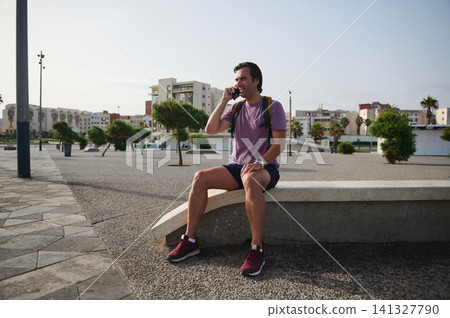Man sitting on city bench talking on phone with backpack in casual summer outfit Man sitting on city bench talking on phone with backpack in casual summer outfit 141327790